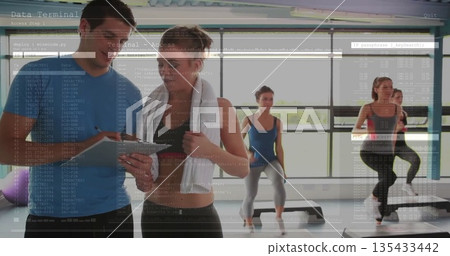 Holding tablet, male instructor guiding trainee in exercise studio with white towel, step platforms 135433442