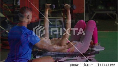 Client wearing pink sports bra bench pressing hex dumbbells at gym, with trainer spotting 135433466