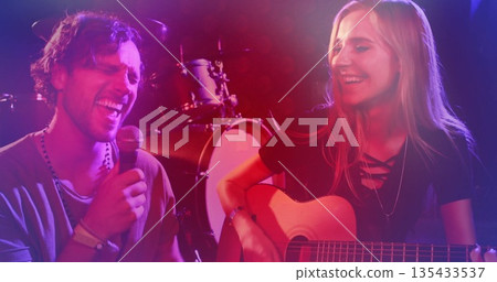 Singing singer holding microphone as guitarist strumming guitar on stage, with drum kit and lights 135433537
