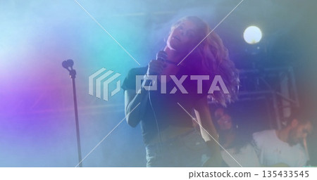 Performing female singer holding handheld microphone on concert stage, with fog and electric guitar 135433545