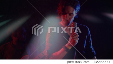Singing singer holding wired microphone on stage, guitarist strumming guitar under spotlights Singing singer holding wired microphone on stage, guitarist strumming guitar under spotlights 135433554