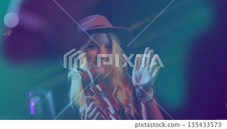 Performing female musician wearing hat holding tambourine while waving on stage, with spotlights 135433573