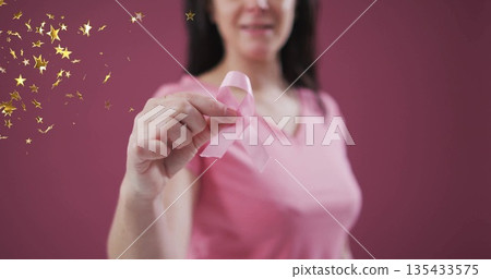Holding pink awareness ribbon, woman wearing pink shirt posing in studio, with gold star confetti 135433575