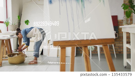 Bending over wicker basket woman wearing denim shirt, apron in art studio, with easel copy space 135433579