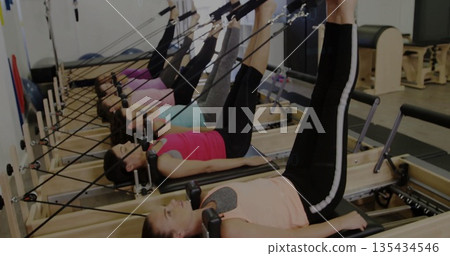 Lifting legs vertically, six women on reformers exercising in pilates studio, with wooden frames Lifting legs vertically, six women on reformers exercising in pilates studio, with wooden frames 135434546