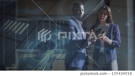 Colleagues in navy blazers examining tablet in boardroom, with holographic data and glass of water 135434558