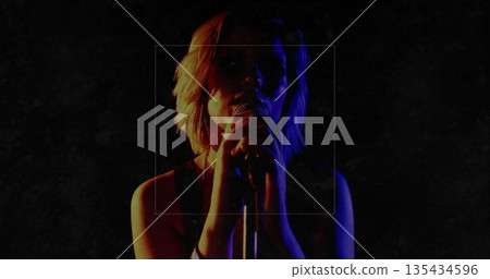 Performing teenage female singer gripping handheld microphone on dark stage, with red blue lighting 135434596