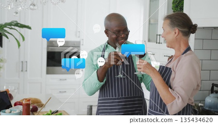 Clinking wine glasses, senior couple in aprons toasting at kitchen island, with digital tablet 135434620
