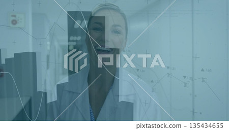 Standing smiling mature woman wearing white lab coat in lab corridor, with floating data charts Standing smiling mature woman wearing white lab coat in lab corridor, with floating data charts 135434655
