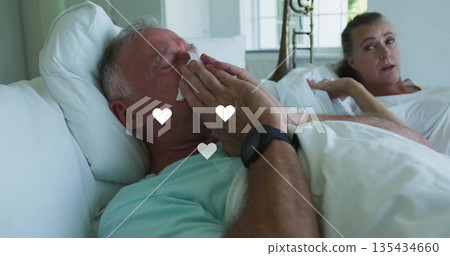 Holding tissue to nose, senior man wearing shirt and wristwatch reclining in bedroom, with pillows Holding tissue to nose, senior man wearing shirt and wristwatch reclining in bedroom, with pillows 135434660