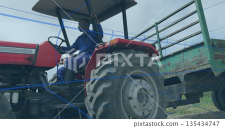 Driving man in blue coveralls steering red tractor on rural field with green trailer, copy space 135434747