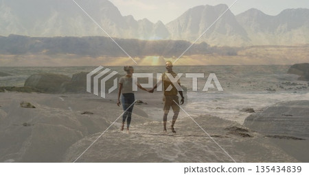 Holding hands couple walking at sandy shoreline with rocks, ocean waves and mountain range 135434839