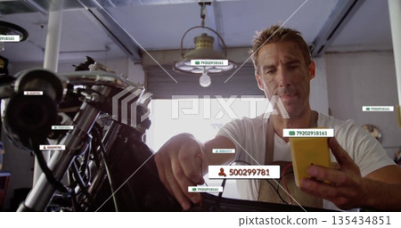 Male mechanic wearing beige apron adjusting motorcycle engine under lamp in garage, with smartphone Male mechanic wearing beige apron adjusting motorcycle engine under lamp in garage, with smartphone 135434851