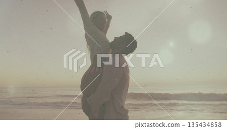 Lifting Hispanic man hoisting White woman high at sandy shoreline, with ocean waves and lens flare 135434858