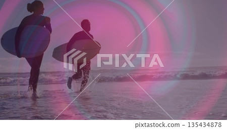 Walking duo of surfers carrying surfboards at sandy beach shoreline, with full-body wetsuits 135434878