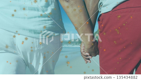 Walking mature couple holding hands and strolling along sandy shoreline, with sparkles overlay 135434882