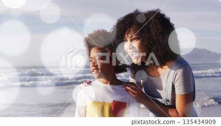 Gazing mother and son sharing moment facing horizon at sandy beach shoreline, with soft lens flares 135434909