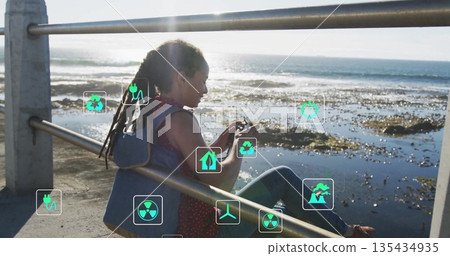 Sitting dreadlocked woman leaning at seaside railing wearing backpack holding phone, with eco icons 135434935