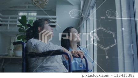 Supporting patient nurse in scrubs holding wheelchair at large window in lounge, with stethoscope 135435392