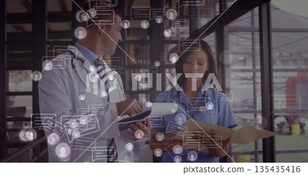 Doctor and nurse reviewing charts in hospital corridor, wearing coat and scrubs with digital icons Doctor and nurse reviewing charts in hospital corridor, wearing coat and scrubs with digital icons 135435416