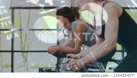 Cycling class participants in sportswear pedaling stationary bikes in studio, with digital overlay 135435477