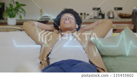 Reclining man holding hands behind head on living room sofa, with wireless earbuds and waveform Reclining man holding hands behind head on living room sofa, with wireless earbuds and waveform 135435602