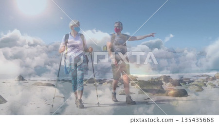 Walking mature couple in hiking attire using trekking poles on shore, with backpacks, face masks Walking mature couple in hiking attire using trekking poles on shore, with backpacks, face masks 135435668