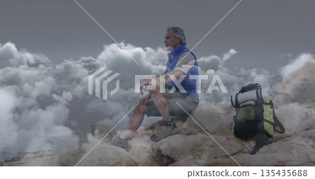 Mature hiker sitting at rocky shoreline overlooking cloudy water, with backpack, vest and boots 135435688