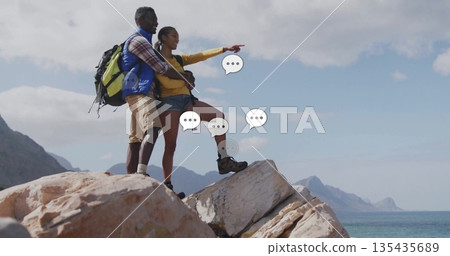 Pointing couple standing on rocky coastal outcrop above ocean, with backpacks and boots, copy space 135435689