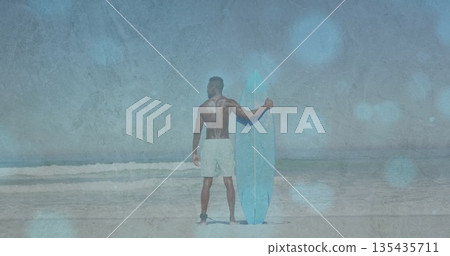 Standing surfer in swim shorts holding light-blue surfboard on sandy shore, preparing for surf Standing surfer in swim shorts holding light-blue surfboard on sandy shore, preparing for surf 135435711