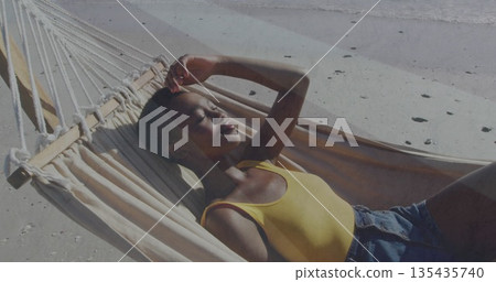 Reclining woman wearing yellow tank top shielding eyes from sun on sandy beach hammock, with waves 135435740