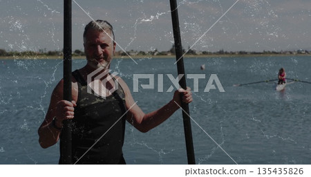 Standing senior male paddler holding two black paddles chest-deep in calm lake, with buoy and scull Standing senior male paddler holding two black paddles chest-deep in calm lake, with buoy and scull 135435826