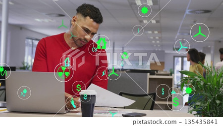 Office worker man reviewing documents at desk in open office, with laptop and floating data icons 135435841