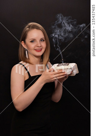 A woman in a black dress blows out a candle on a bento cake. Smoke on a black background. A girl's birthday, happiness, a gift. High quality photo A woman in a black dress blows out a candle on a bento cake. Smoke on a black background. A girl's birthday, happiness, a gift. High quality photo 135437485