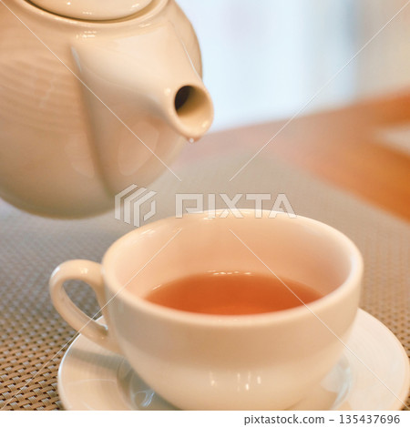 Warm cup of tea being poured from white teapot into a cup on a saucer in a cozy Warm cup of tea being poured from white teapot into a cup on a saucer in a cozy 135437696