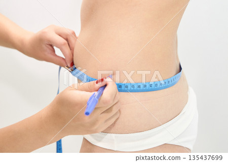 Close-up of female measuring waist with tape for fitness and health monitoring Close-up of female measuring waist with tape for fitness and health monitoring 135437699