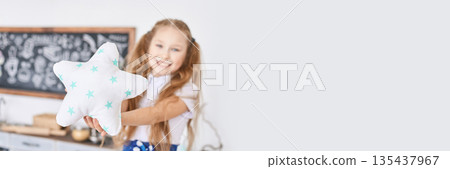 Smiling caucasian young girl holding star pillow in bright room with chalkboard Smiling caucasian young girl holding star pillow in bright room with chalkboard 135437967