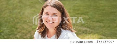 Smiling young caucasian female sitting on grass in sunny outdoor environment Smiling young caucasian female sitting on grass in sunny outdoor environment 135437997
