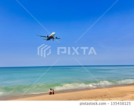 A young happy couple is sitting on the beach, and a plane is flying in the blue sky A young happy couple is sitting on the beach, and a plane is flying in the blue sky 135438125