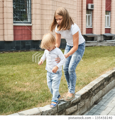 Caucasian female teen helping blonde young child walk on curb in urban park 135438140