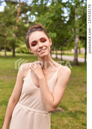 Young caucasian female in cream dress with bold makeup smiling in park setting Young caucasian female in cream dress with bold makeup smiling in park setting 135438176