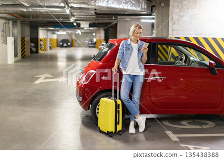 A woman stands in a parking garage with her luggage, looking at her phone next to a red car 135438388
