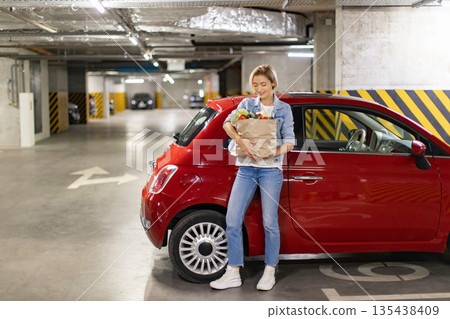 A woman smiles while holding a bag of groceries next to her red car in a parking garage 135438409