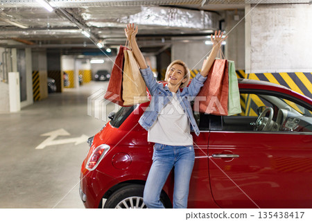 A woman stands next to her car in a parking garage, holding shopping bags and celebrating her purchases A woman stands next to her car in a parking garage, holding shopping bags and celebrating her purchases 135438417