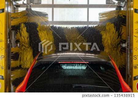 Red vehicle being cleaned in an automatic brush car wash with water and foam. Red vehicle being cleaned in an automatic brush car wash with water and foam. 135438915