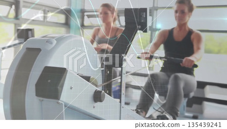 Rowing athletic friends pushing fluid strokes on stationary machines at gym, with digital monitors 135439241