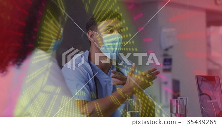 Gesturing doctor in scrubs and mask, speaking in consultation room with laptop and digital overlay 135439265
