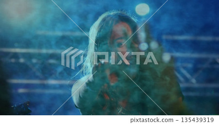 Pointing female singer standing amid haze under blue lights on concert stage, with mic and truss 135439319