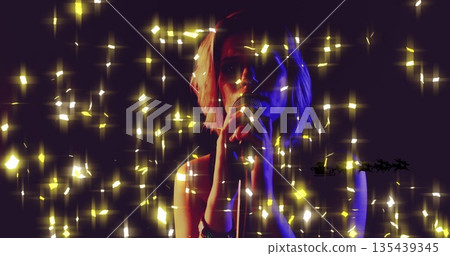 Singing teenage girl wearing sleeveless top on stage, holding microphone amid golden sparkles 135439345
