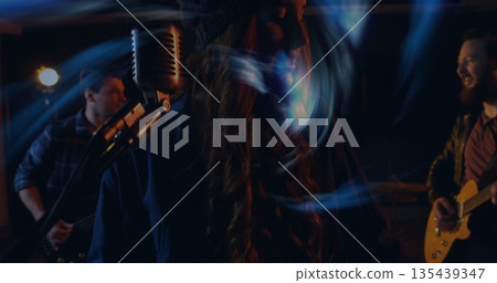 Performing female singer singing into vintage mic on stage, with bassist, guitarist under lights 135439347